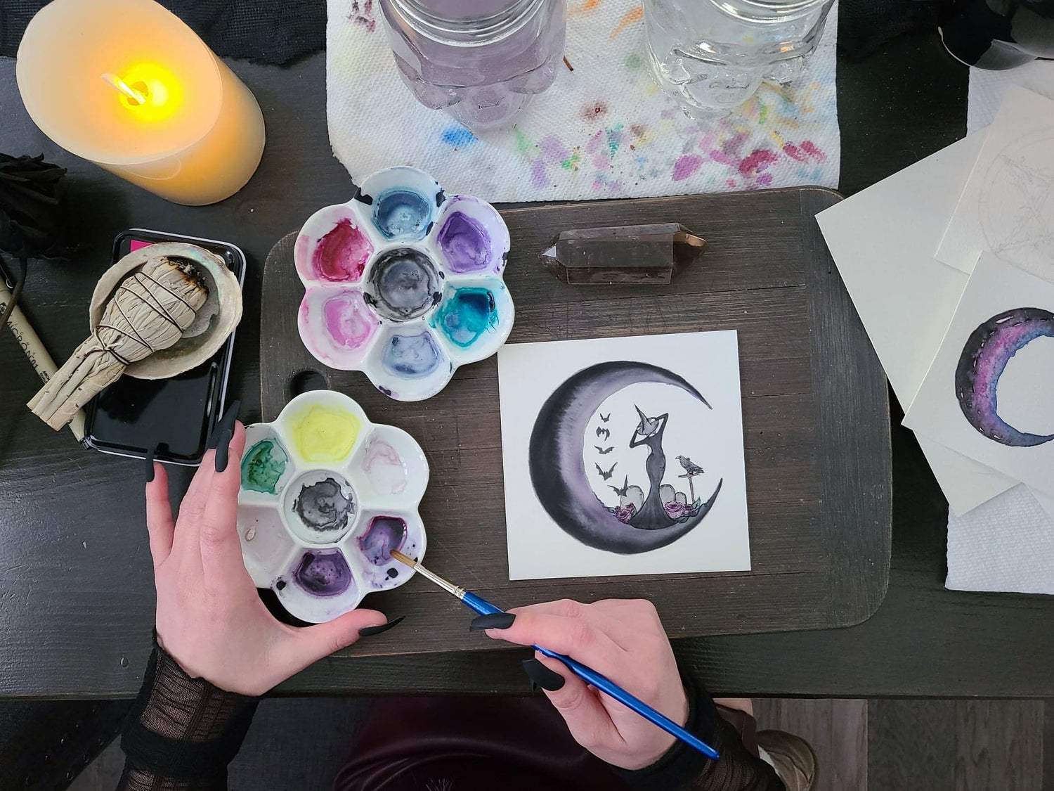 Watercolor artist holding paint with a moon and witchy woman on the canvas. Magic items and art supplies surround the studio space.